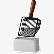 Black wire cat litter scoop with wooden handle standing in white ceramic holder base