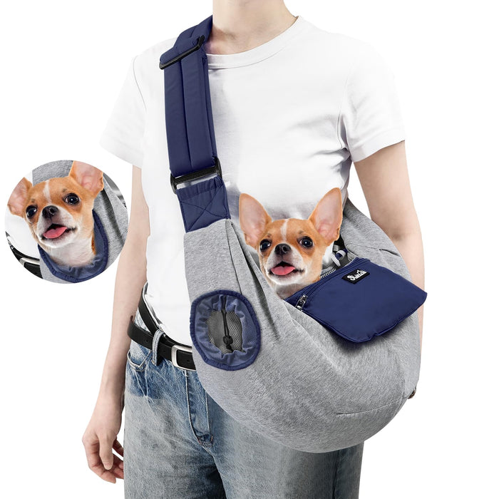 Person wearing gray dog sling carrier with navy accents, small chihuahua inside with tongue out