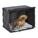 Small tan dog sitting inside black wire crate with dark cover, white bedding and metal bowls
