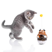 Gray tabby kitten reaching toward brown cow-shaped treat dispenser toy with yellow ball on top