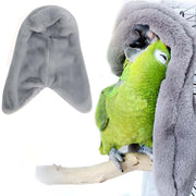 Green parrot wearing gray fleece corner bird blanket hood while perched on person's hand