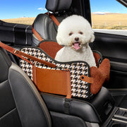 White fluffy dog sitting in brown houndstooth patterned car seat with safety straps in vehicle
