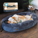 Woman and golden retriever cuddling together in large gray plush oval dog bed next to product box