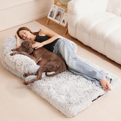 Woman lying on gray fluffy orthopedic dog bed with brown dog, relaxing on wooden floor
