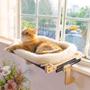 Orange tabby cat relaxing on wooden window perch with white fleece cushion by sunny window